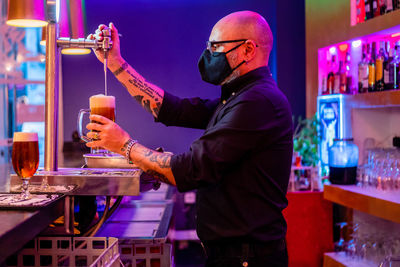 Side view of serious barkeeper in eyeglasses pouring beer into glass while working in bar during coronavirus pandemic