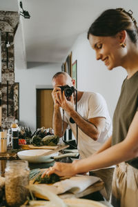 Colleague photographing female chef through camera while preparing food in studio