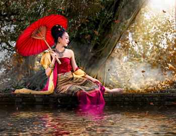 Young woman with umbrella sitting by lake in forest