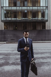 Male entrepreneur with bag using smart phone while standing in city