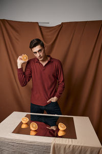 Portrait of young man holding orange standing against brown background