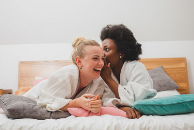 Woman whispering friend while sitting on bed