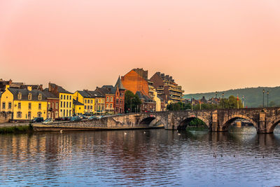 Bridge over river by buildings against sky during sunset