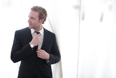 Thoughtful businessman adjusting necktie while standing amidst white curtains