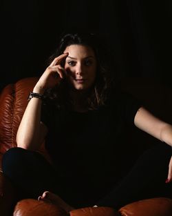 Portrait of young woman touching forehead while sitting against black background
