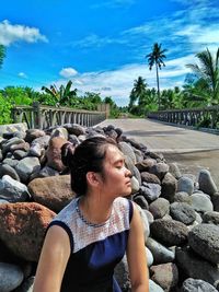 Young woman sitting on retaining wall by beach against sky