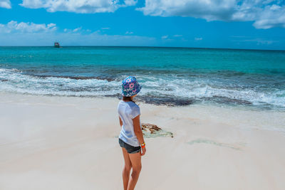 Rear view of girl wearing hat while standing at beach against cloudy sky