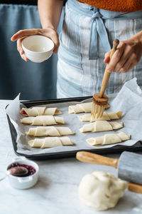 From above crop cook holding white bowl and diligently brushing tasty croissants on baking sheet on table in kitchen
