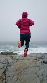 Rear view of person jumping over rocks by sea against sky