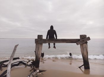 Rear view of man sitting on wooden structure while looking at sea