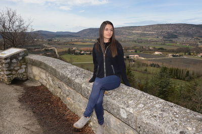 Full length of young woman looking away while sitting on observation point against sky