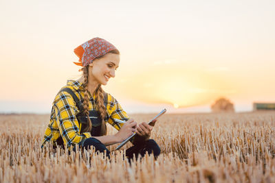 Smiling woman writing on clipboard while sitting in farm during sunset