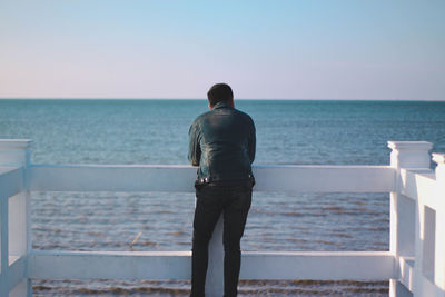 Rear view of young man looking at sea while standing by railing against clear sky