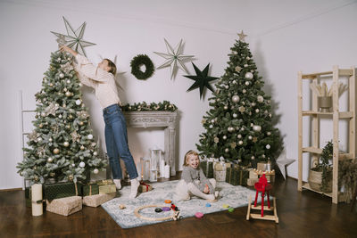 Mother decorating christmas tree with cute daughter sitting on carpet at home