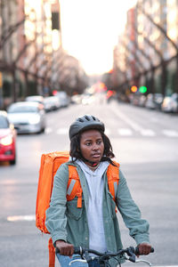 Serious african american female in helmet with orange thermo backpack sitting on bicycle near road while delivering order