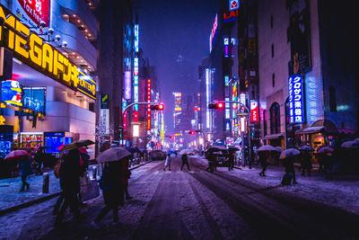 People walking on illuminated street amidst buildings during snowfall