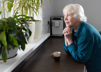 An attractive elderly woman drinks espresso in the kitchen and looks out the window