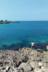 High angle view of man with arms outstretched standing at rocky beach against clear blue sky