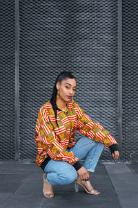 Full body young ethnic female in trendy outfit with long braid sitting on haunches on pavement and looking at camera against gray wall on city street