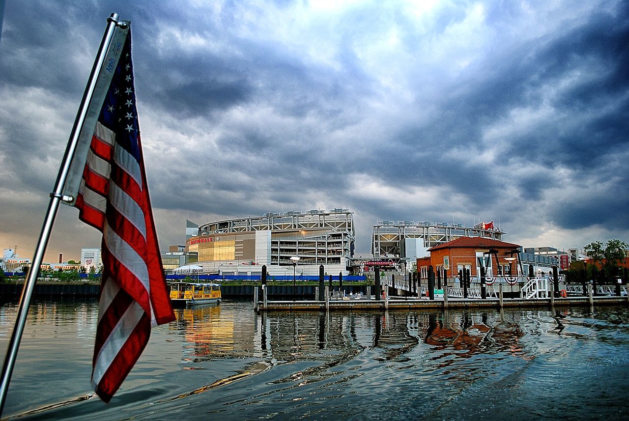 building exterior, architecture, built structure, water, cloud, city, waterfront, flag, sky, rippled, sea, blue, outdoors, development, skyscraper, day, cloud - sky, multi colored, national flag, modern, cloudy, no people, water surface, building story, office building