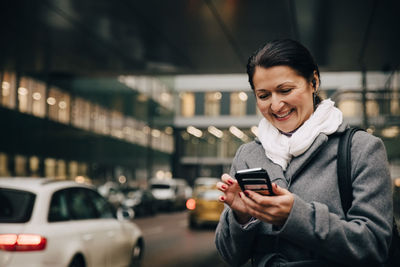 Smiling businesswoman using smart phone while standing on city street against building