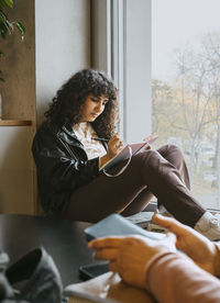 Young woman writing in book while sitting by window at university