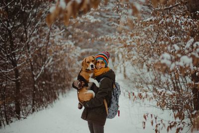 Portrait of smiling young woman carrying dog while standing in forest during winter