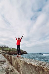 Woman standing with arms outstretched on retaining wall by sea against sky