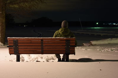 Rear view of man sitting on bench during winter at night