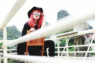 Portrait of young woman wearing sunglasses while sitting on chair by railing