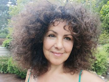 Close-up of smiling mature woman with curly brown hair against trees