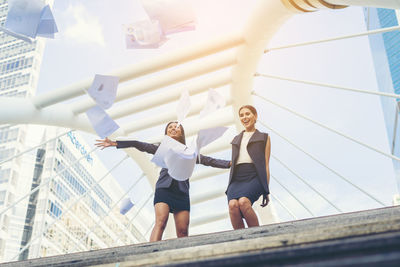 Low angle view of happy businesswomen throwing papers against buildings in city 