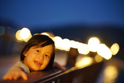 Portrait of girl smiling while leaning on railing against illuminated lights at dusk