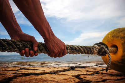 Cropped hands of man holding rope against sky