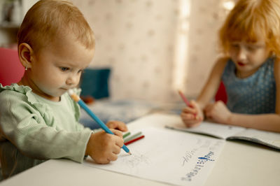 Cute baby girl drawing on paper with sister doing homework at table