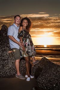 Full length portrait of young couple standing on shore at beach against sky during sunset