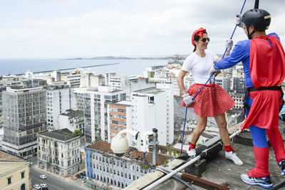 Woman wearing hero costume being prepared by a man to descend a tall rappel building. 