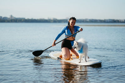 Cheerful woman with dreadlocks paddleboarding with her dog snow-white japanese spitz on sup board