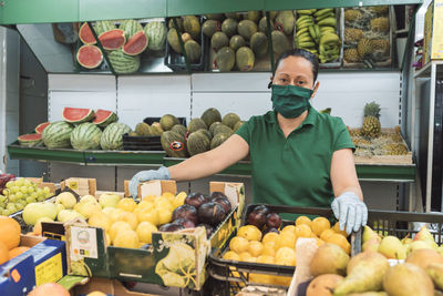 A shopkeeper with a mask poses in front of her fruit shop