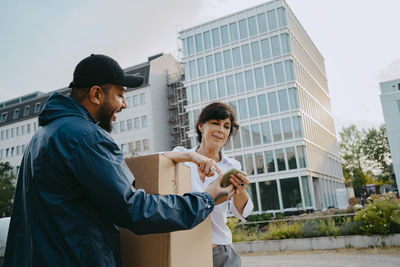 Mature woman doing digital signature while receiving parcel from male delivery person in front of building