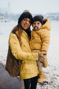 Portrait of smiling mother carrying son o footpath during winter