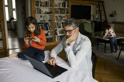Freelancer working on laptop by daughter using smart phone on table while girl playing in background