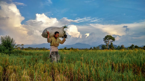 Farmer carrying sack of crops on field against sky