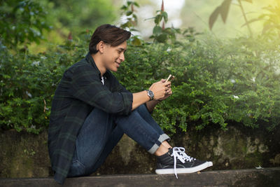 Full length of young man using phone while sitting by plants