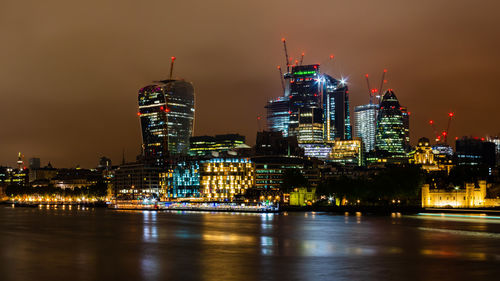 Illuminated buildings by river against sky at night