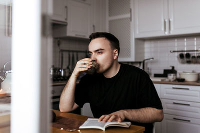 Young man drinking tea and reading a book in his modern light kitchen