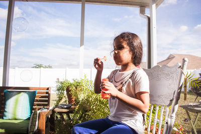 Cute girl blowing bubble while sitting on chair