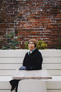 Portrait of senior woman wearing warm clothing sitting on bench against brick wall in city