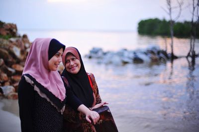 Smiling mother with daughter in hijab standing at beach against sky during sunset