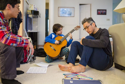 A small boy plays guitar for his father and uncle in living room